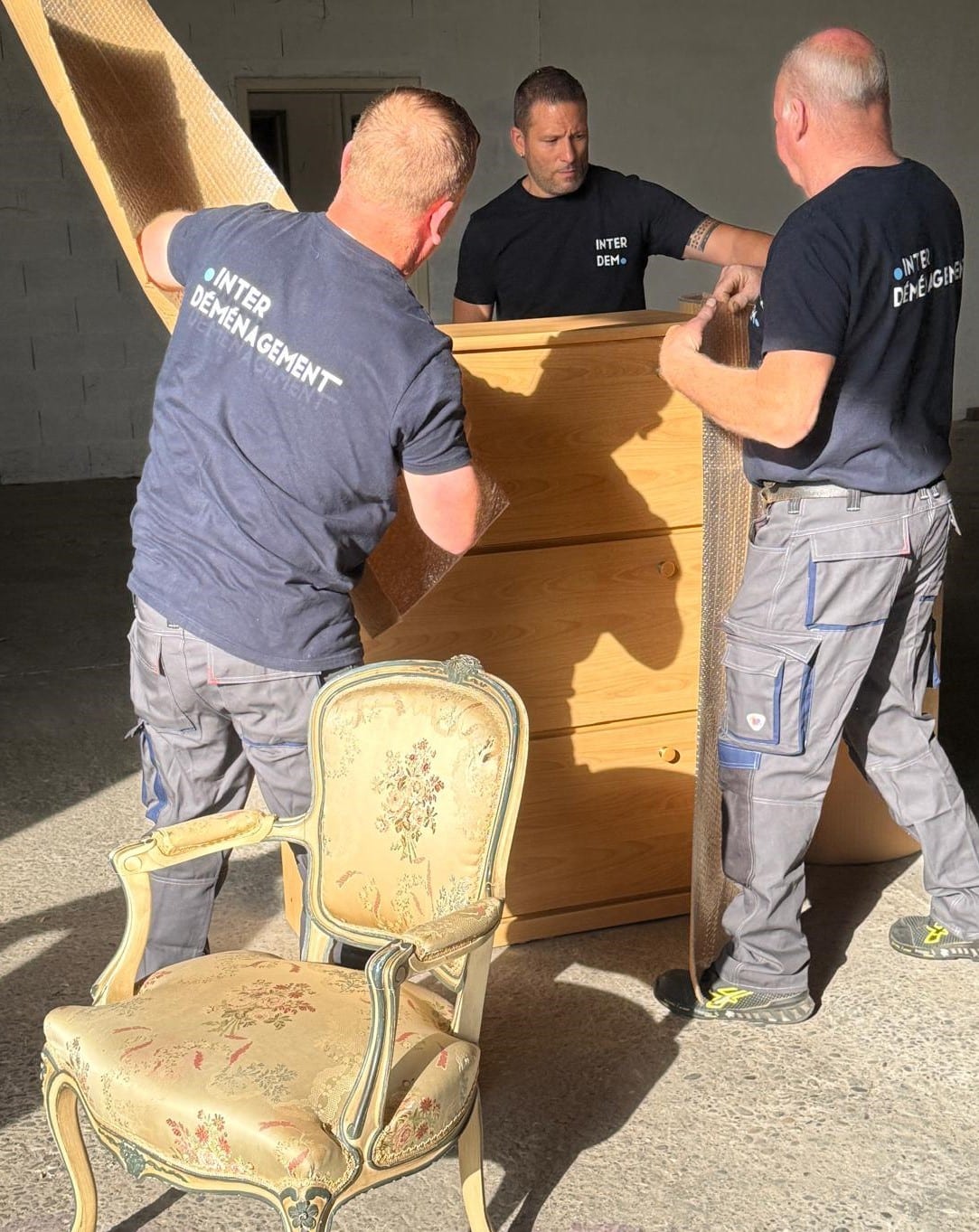 Trois déménageurs d’Inter Déménagement en train de couvrir un meuble avec une couverture de protection, illustrant le soin apporté à la préparation avant transport.